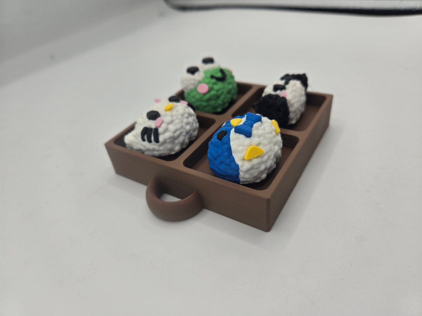 Angled view of the Sanrio themed rice ball tray clicker fidget showing the tray depth and keychain loop, four rice ball characters, 3D printed