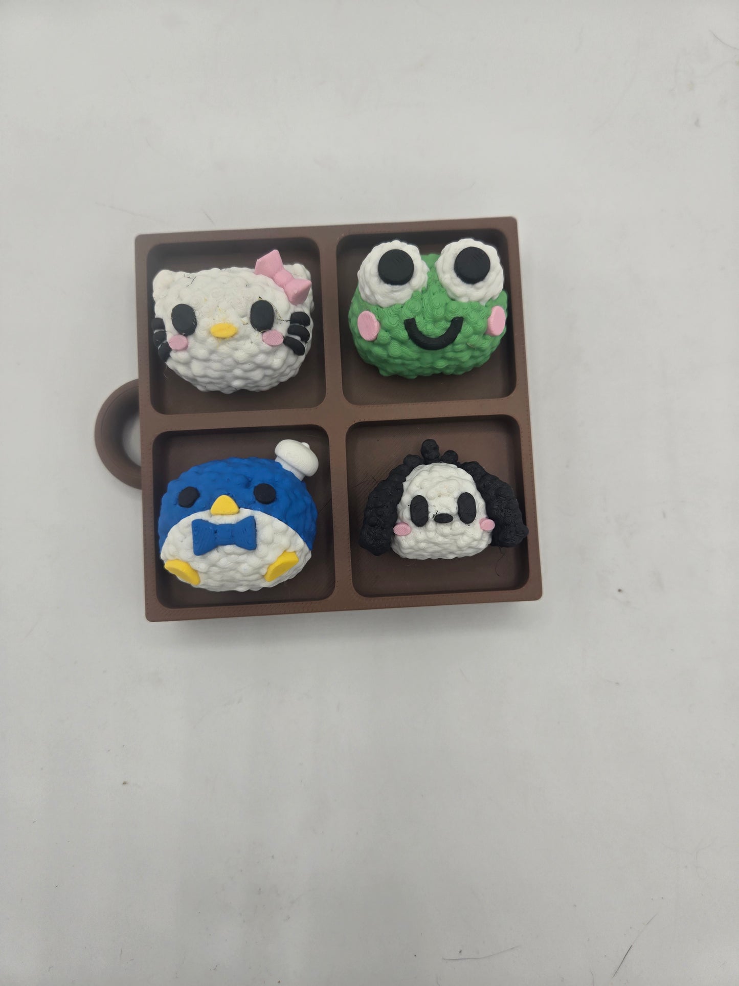 Sanrio themed rice ball tray clicker fidget from above — kitty, frog, penguin, and puppy rice balls in a brown bento-style tray with keychain loop, 3D printed