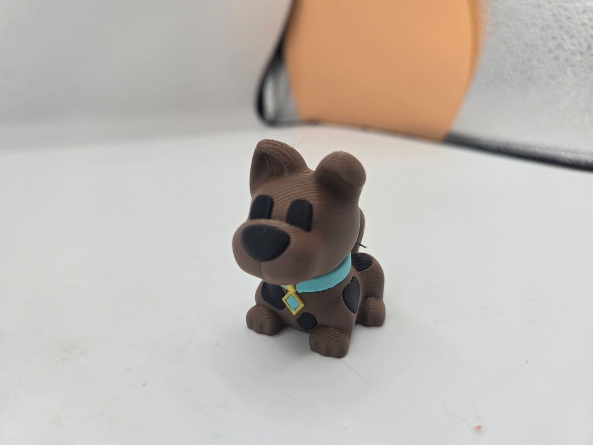 Mini Scooby Doo inspired dog figurine 3D print in brown with black spots and a teal collar, front view on desk
