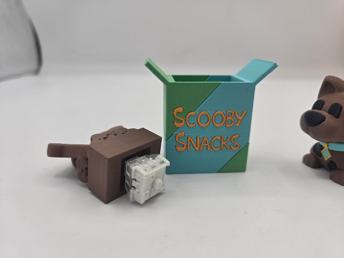 Scooby Snacks key clicker 3D print tipped over to show the mechanical keyboard switch inside the base, with box and dog figurine