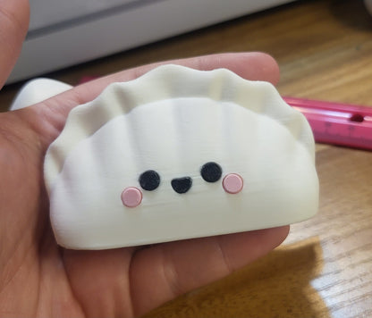 3D printed dumpling figure held in a hand for size reference — white kawaii dumpling with pink cheeks, about 2 inches tall