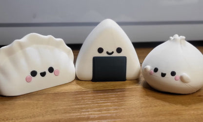 Kawaii trio lineup — 3D printed dumpling, onigiri, and bao figures side by side on a wooden desk; the bao is available in a separate listing