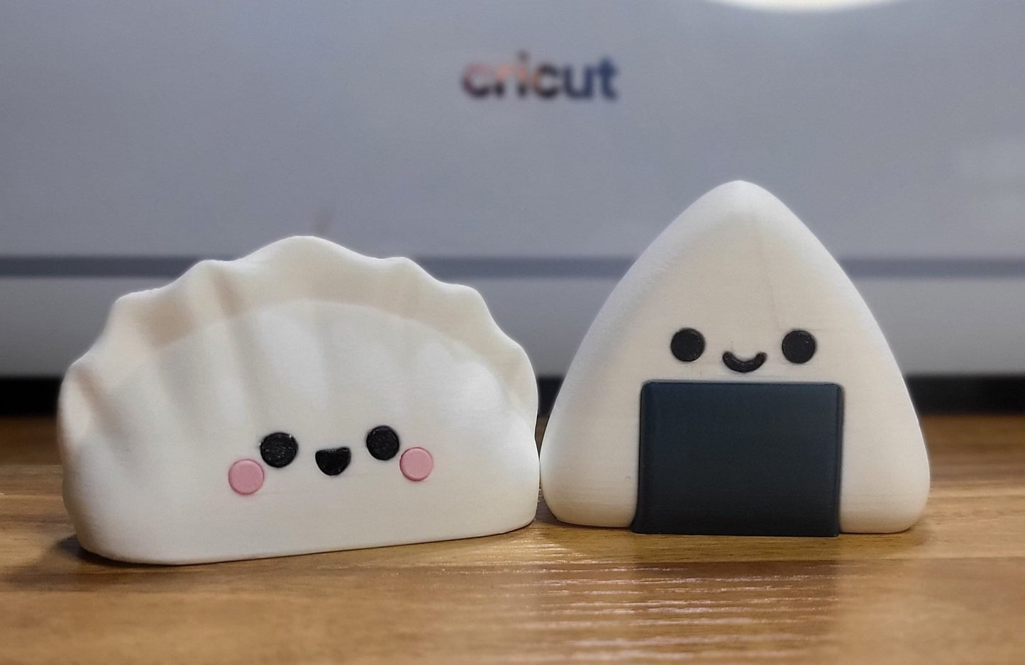 Dumpling and Onigiri 3D printed kawaii figures side by side on a wooden desk — smiling white dumpling with pink cheeks next to a white onigiri with black seaweed wrap