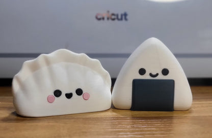 Dumpling and Onigiri 3D printed kawaii figures side by side on a wooden desk — smiling white dumpling with pink cheeks next to a white onigiri with black seaweed wrap
