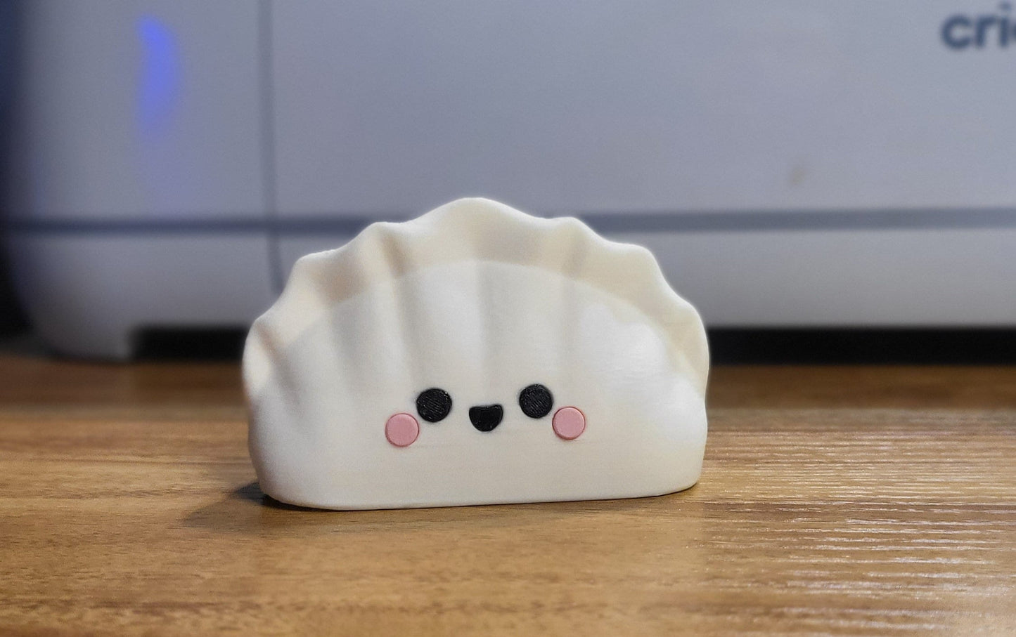Kawaii dumpling figure front view — 3D printed in white filament with a smiling face and pink cheeks, pleated top, standing on a wooden desk
