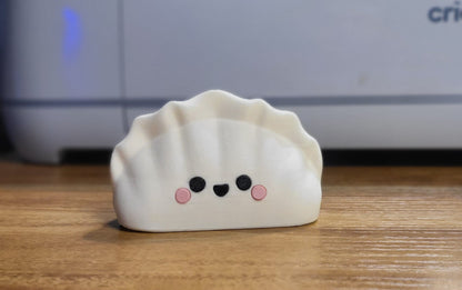 Kawaii dumpling figure front view — 3D printed in white filament with a smiling face and pink cheeks, pleated top, standing on a wooden desk