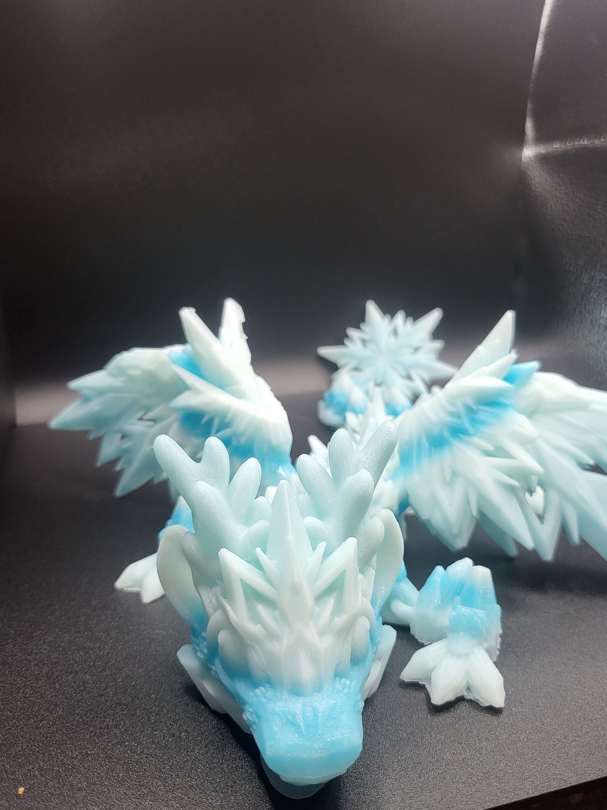Baby Winter Dragon facing the camera, antlers and feathered crystal wings spread — 3D printed in frosty white-blue gradient with a snowflake ornament behind