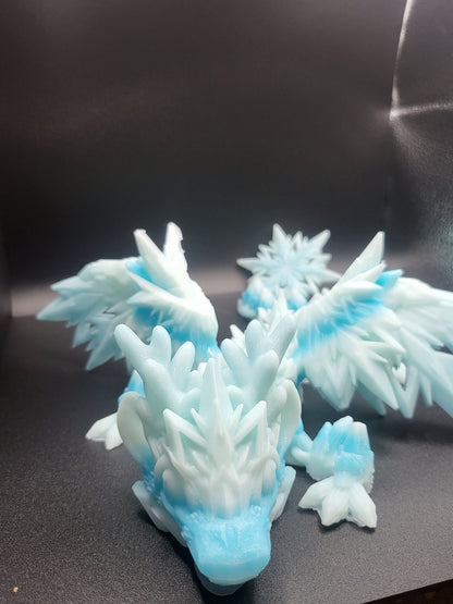 Baby Winter Dragon facing the camera, antlers and feathered crystal wings spread — 3D printed in frosty white-blue gradient with a snowflake ornament behind