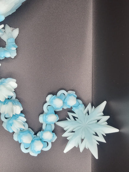Close-up of the Baby Winter Dragon's articulated tail segments curled beside a 3D printed snowflake star — icy blue and white filament