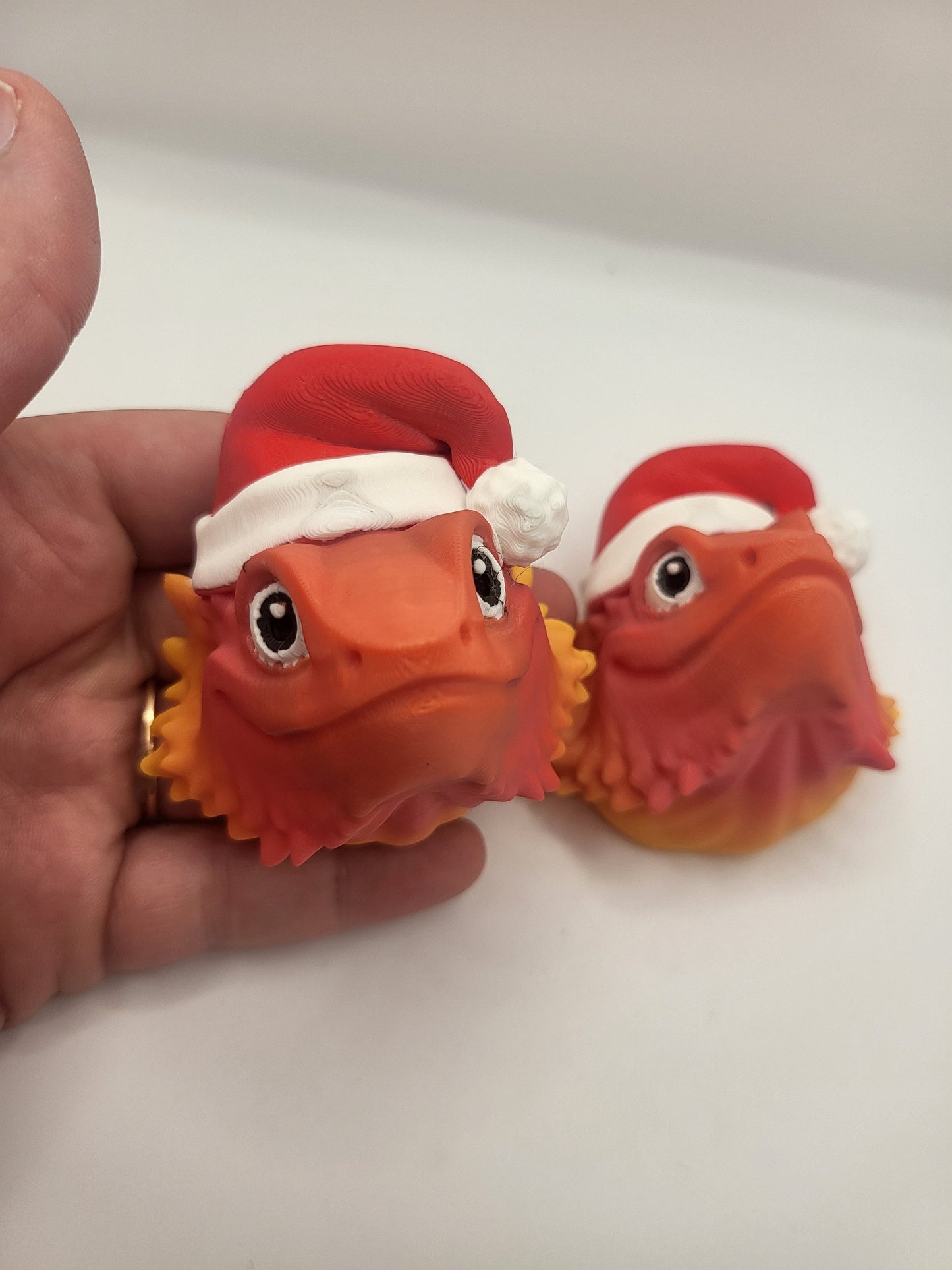 Two sizes of the 3D printed Bearded Dragon with Santa Hat held in a hand — orange-to-yellow gradient faces with red and white Santa hats