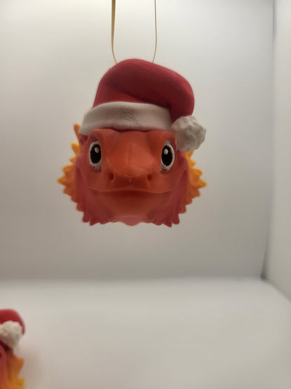 Bearded Dragon with Santa Hat ornament hanging from a gold ribbon, front view — cute wide-eyed orange bearded dragon face, 3D printed