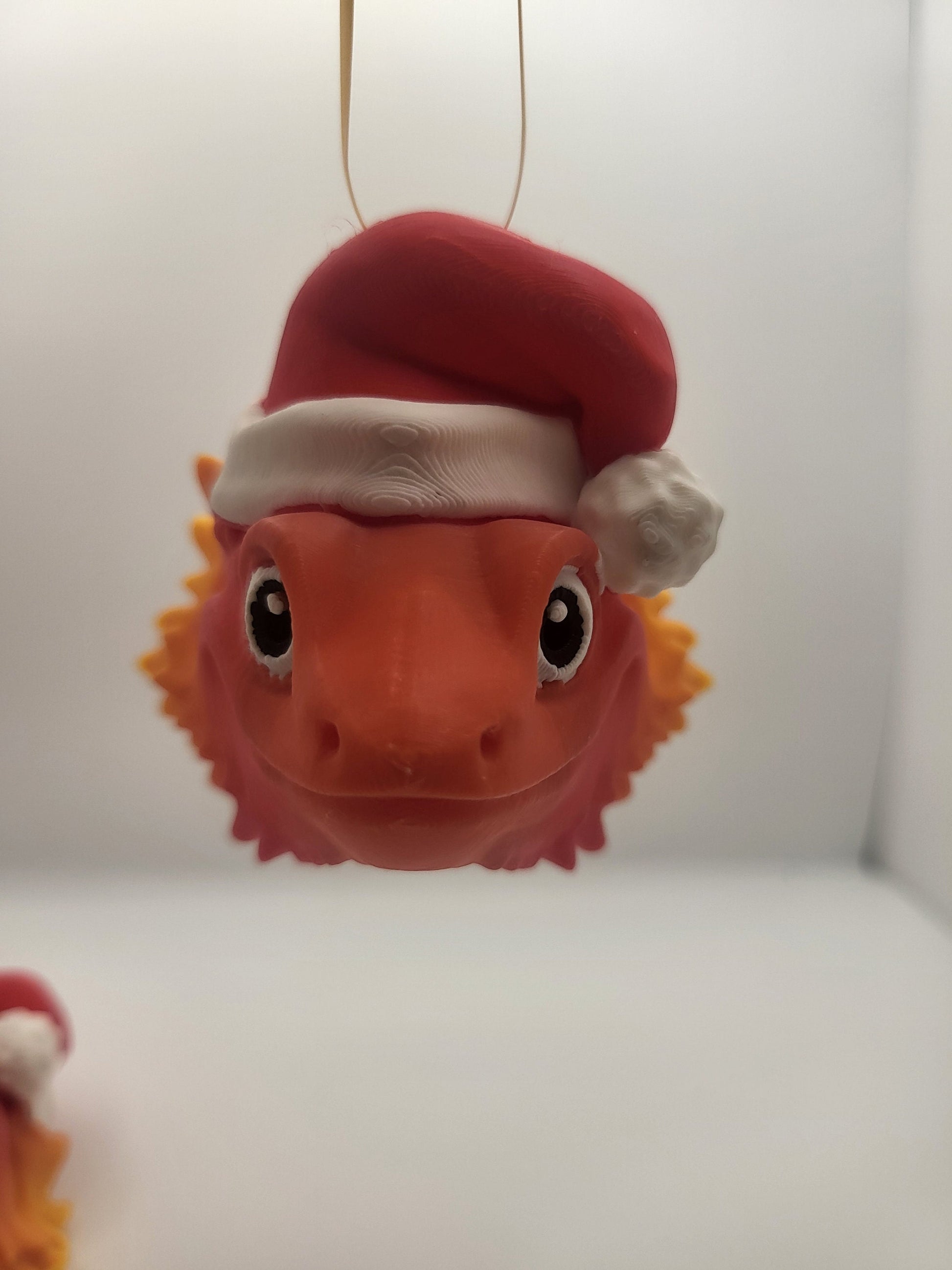 Close-up of the hanging Bearded Dragon with Santa Hat ornament — big friendly eyes, spiky orange beard, and white pom-pom hat, 3D printed
