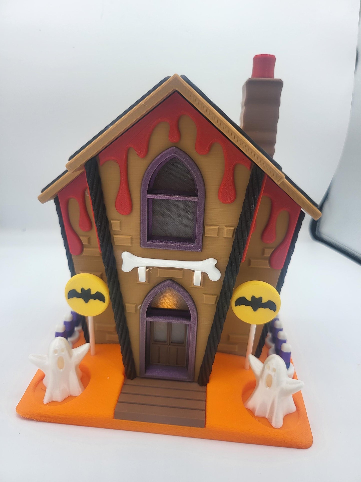 Halloween Gingerbread House front view — purple arched windows, bone sign above the door, bat lollipop signs, and ghost figurines flanking the steps, 3D printed