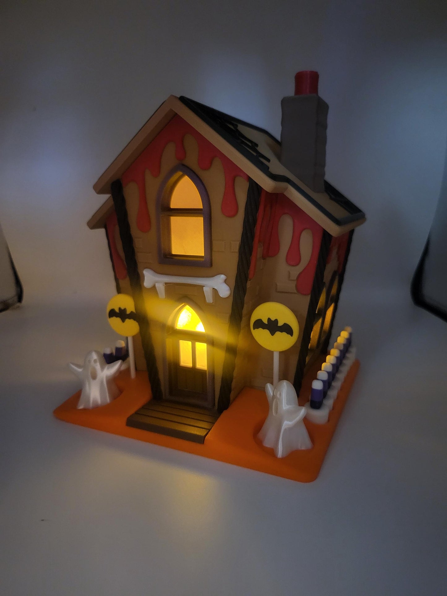 Halloween Gingerbread House lit at night — 3D printed house glowing from within, red ooze roof drips, ghosts, bat lollipop signs, and candy corn fence on the orange base