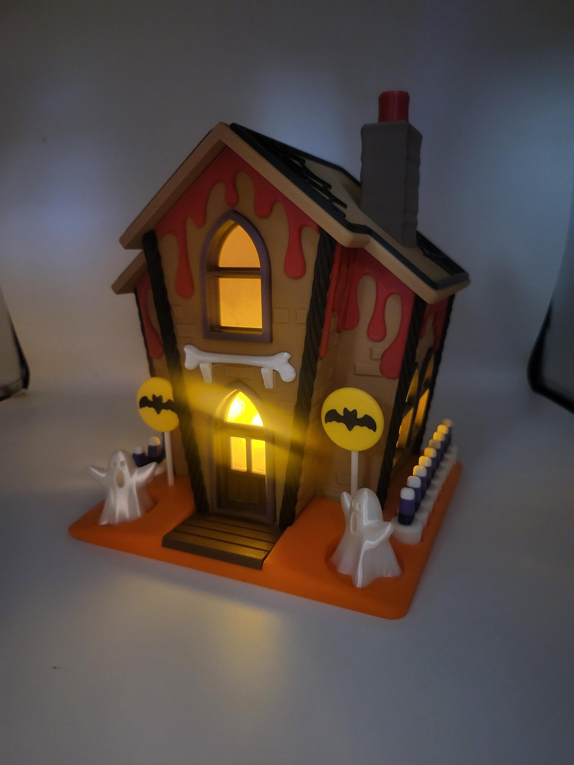 Halloween Gingerbread House lit at night — 3D printed house glowing from within, red ooze roof drips, ghosts, bat lollipop signs, and candy corn fence on the orange base