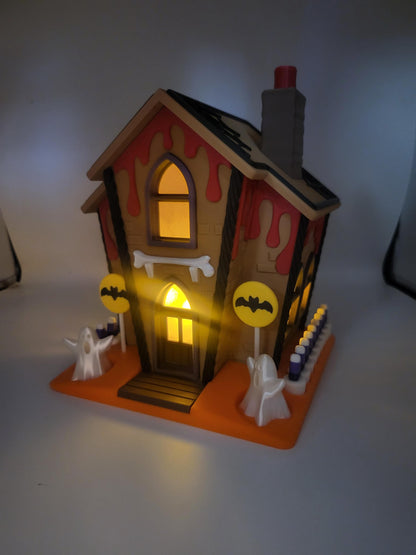 Halloween Gingerbread House lit at night — 3D printed house glowing from within, red ooze roof drips, ghosts, bat lollipop signs, and candy corn fence on the orange base