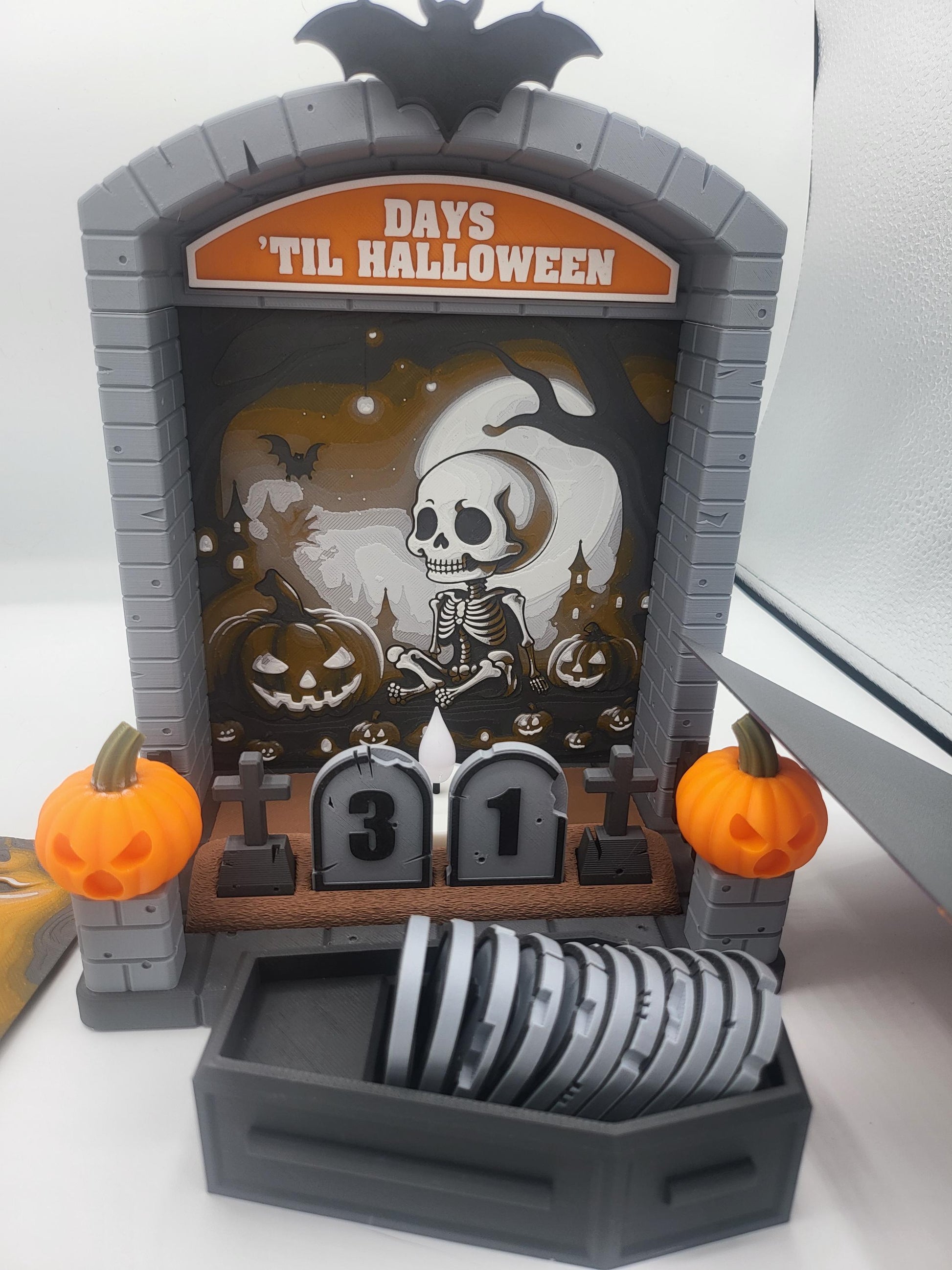 Halloween Countdown with skeleton graveyard backdrop in daylight — coffin storage filled with tombstone-shaped numbers in front of the display, 3D printed