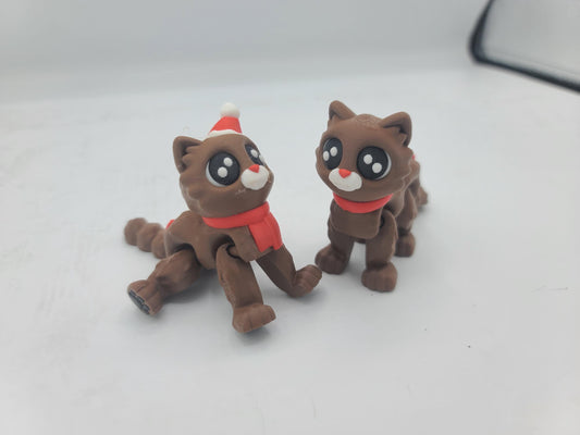 Two Cocoa Kitty flexi cats in a playful crouch — chocolate-brown 3D printed cats, one in a Santa hat with red scarf, one with a red scarf only