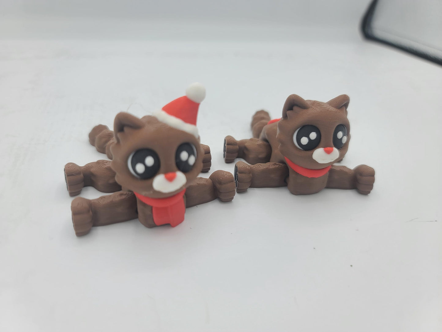Two Cocoa Kitty flexi cats lying flat with legs stretched out, showing their articulated bodies — Santa Hat and scarf styles, 3D printed