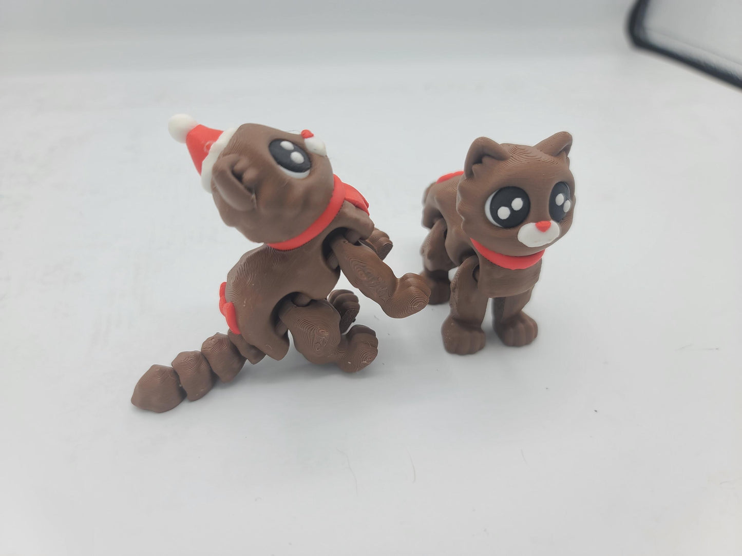 Side view of the Cocoa Kitties at play — Santa Hat kitty rearing up with segmented tail curled while the No Hat kitty stands nearby, 3D printed