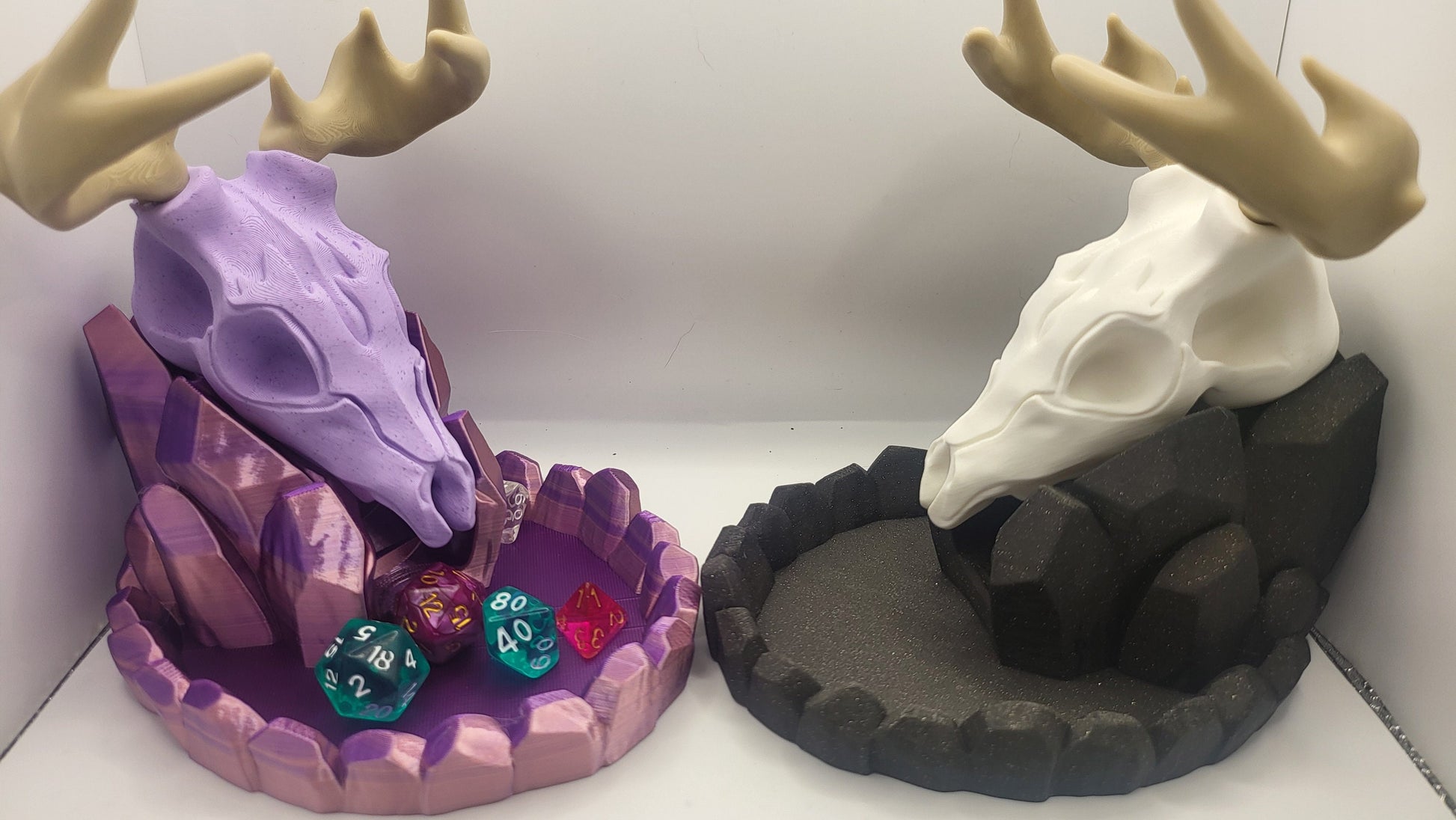 May include: Two 3D printed dice trays, one purple and one black, shaped like a deer skull with antlers. The purple tray has a crystal-like base and is filled with dice. The black tray has a rocky base and is empty.
