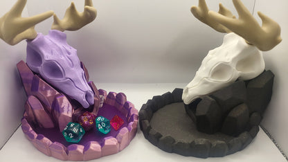 May include: Two 3D printed dice trays, one purple and one black, shaped like a deer skull with antlers. The purple tray has a crystal-like base and is filled with dice. The black tray has a rocky base and is empty.