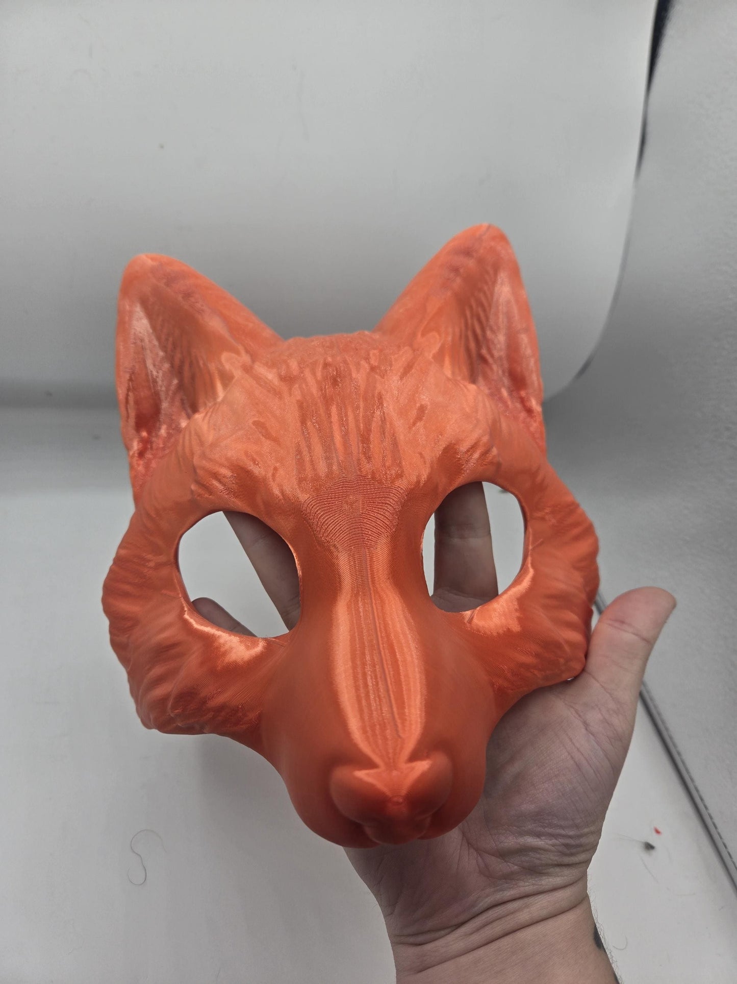 May include: An orange fox mask, likely 3D printed, held in a hand. The mask features pointed ears, eye holes, and a prominent nose. The surface shows visible layer lines from the printing process, giving it a textured appearance. The mask is a solid, uniform orange color.
