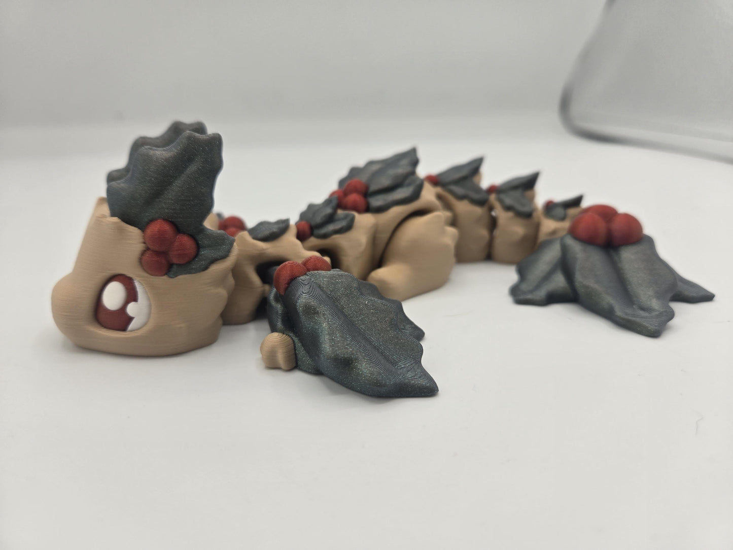 Mistletoe Dragon flexi profile view — cartoon eye, dark holly leaf crest and fanned tail on a beige articulated body, 3D printed