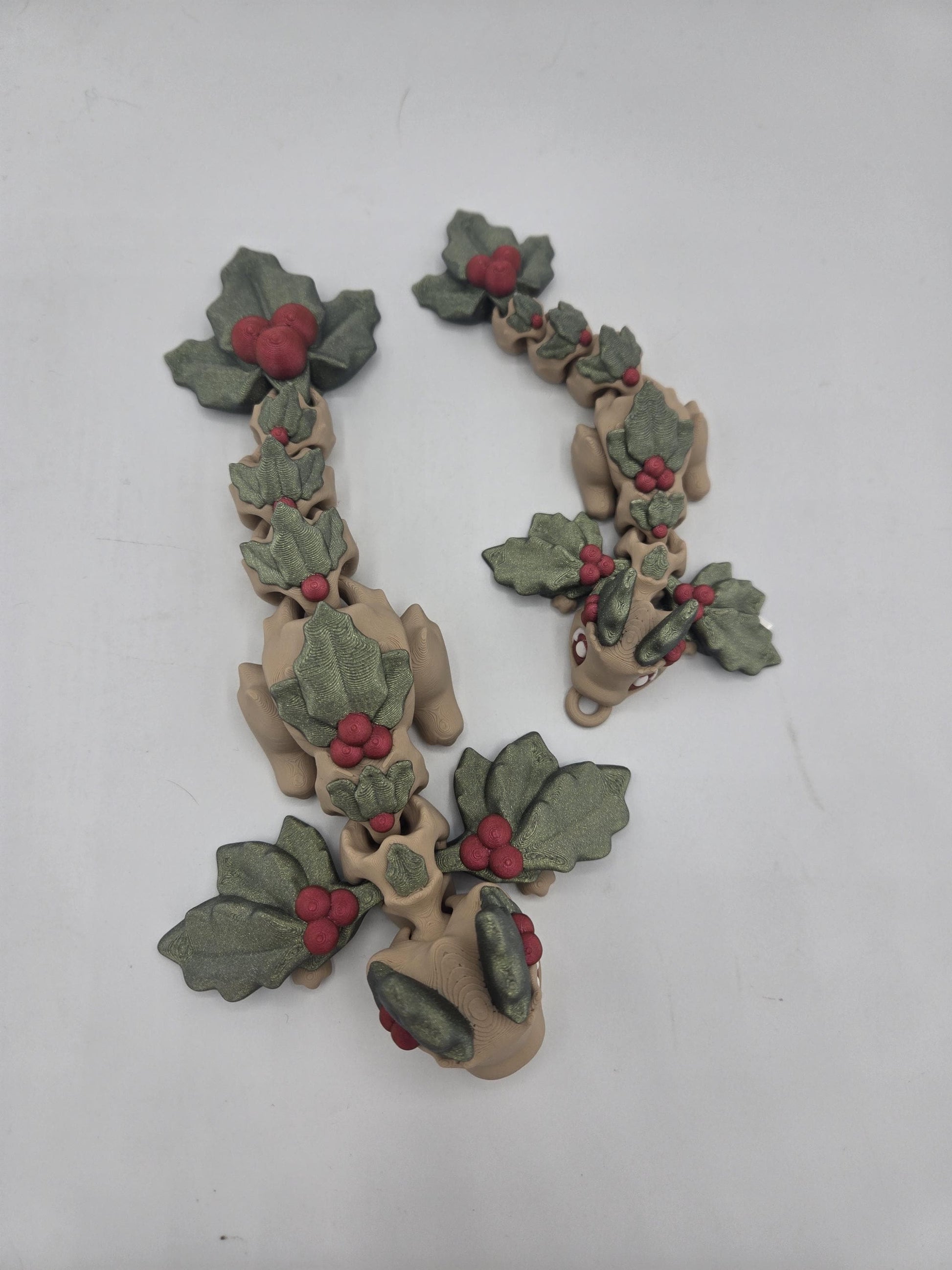 Overhead view of full size and keychain Mistletoe Dragon flexis — beige 3D printed dragons covered in holly leaves and red berries