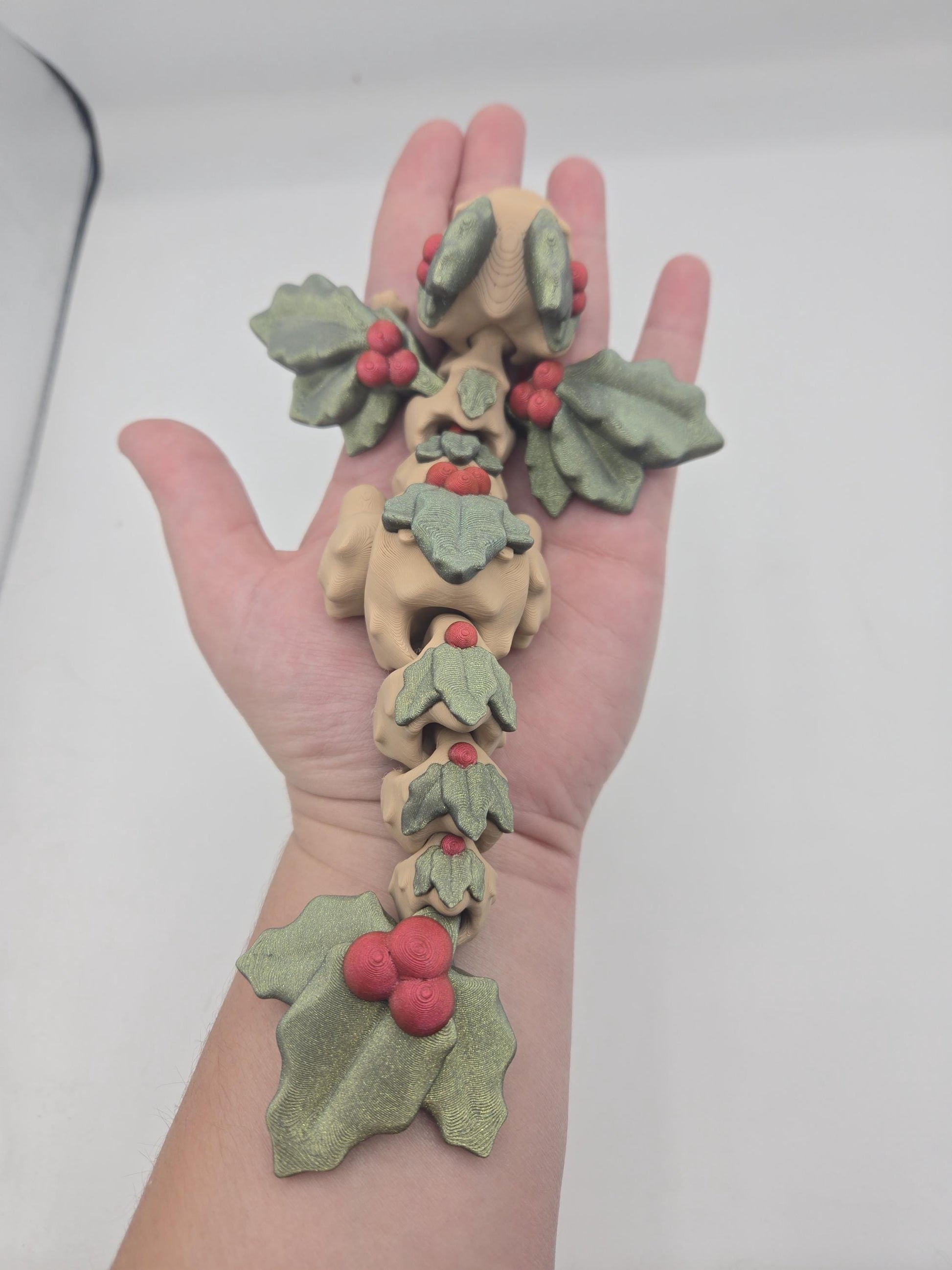 Mistletoe Dragon flexi draped along a hand and forearm — flexible articulated 3D print with green holly leaves and red berry clusters