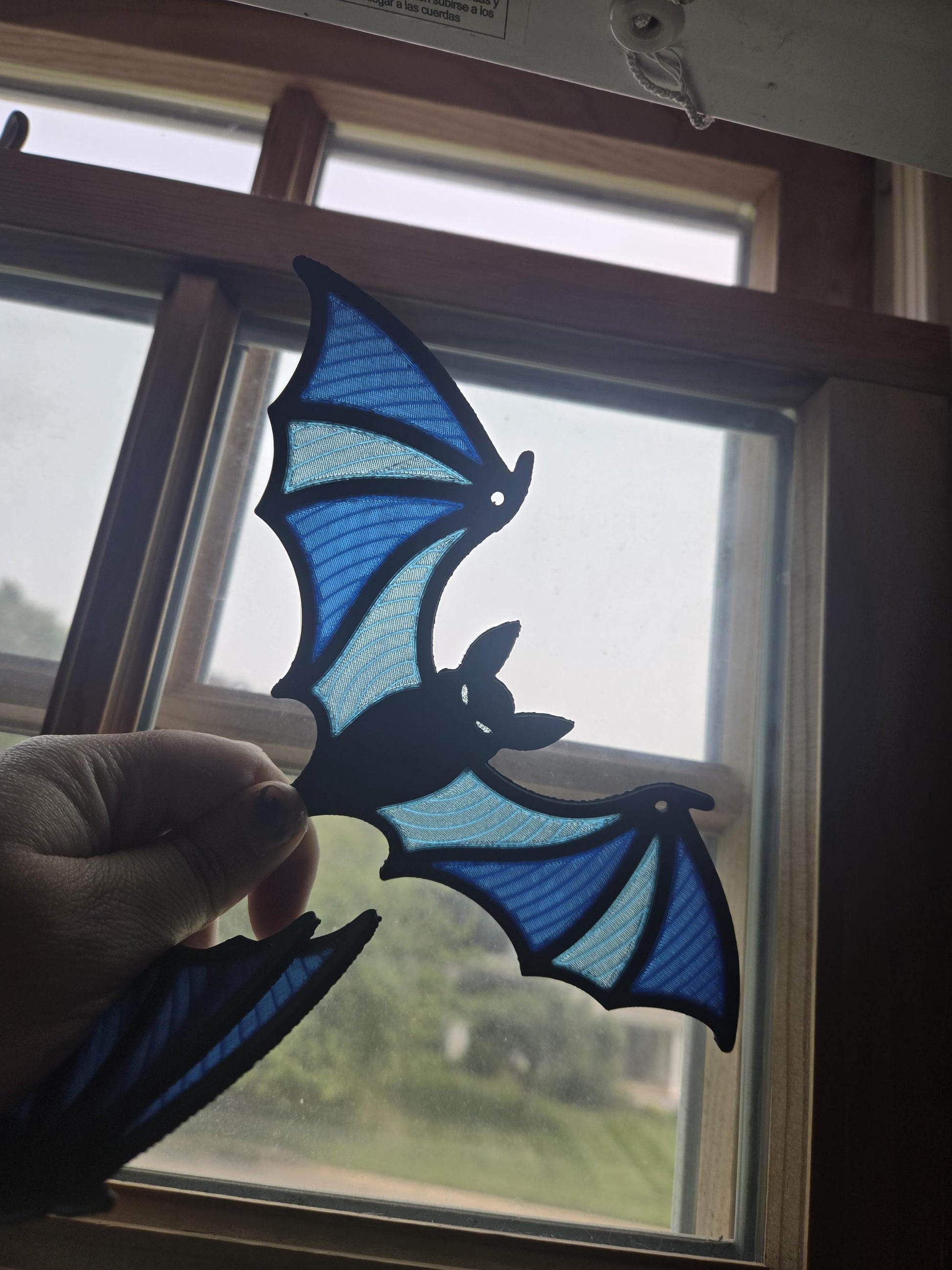 Spooky bat stained glass style window hanging held up to a window — translucent blue wing panels glowing in daylight, 3D printed