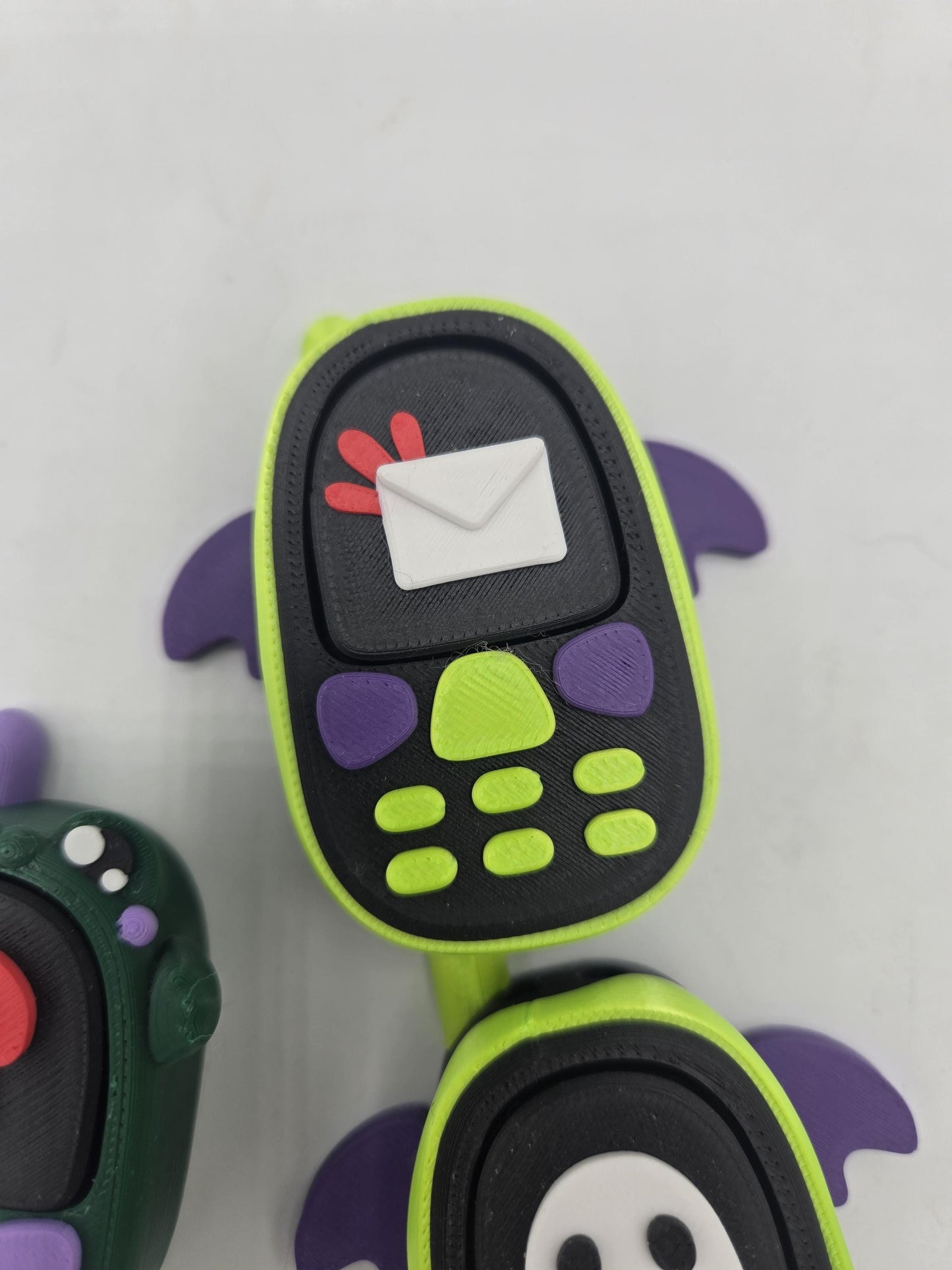 Kawaii bat phone clicker with envelope screen design — lime green with purple bat wings, ghost bat and frog phones nearby, 3D printed