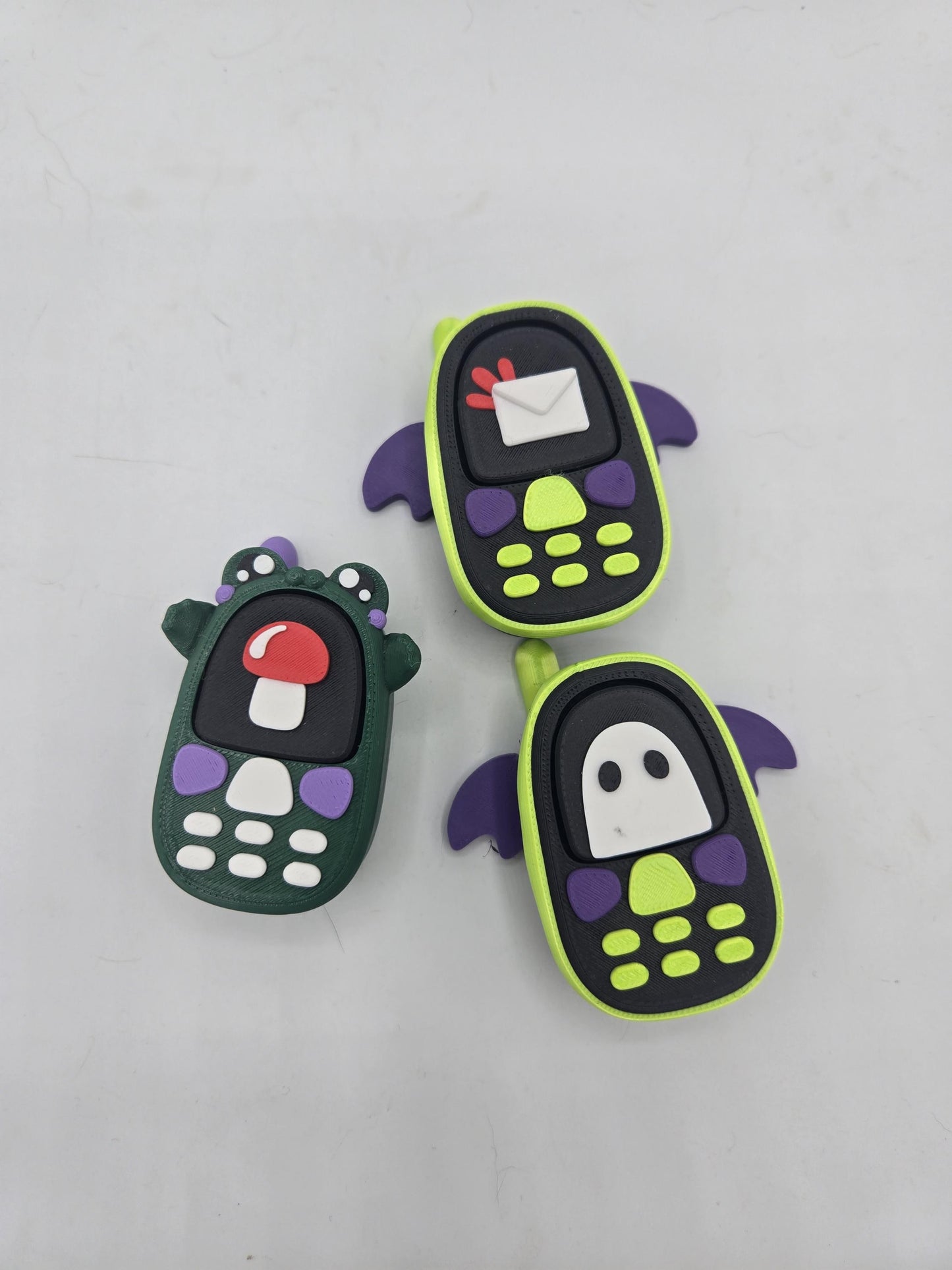 All three kawaii phone clicker styles together — frog with mushroom screen, bat with envelope screen, and bat with ghost screen, 3D printed
