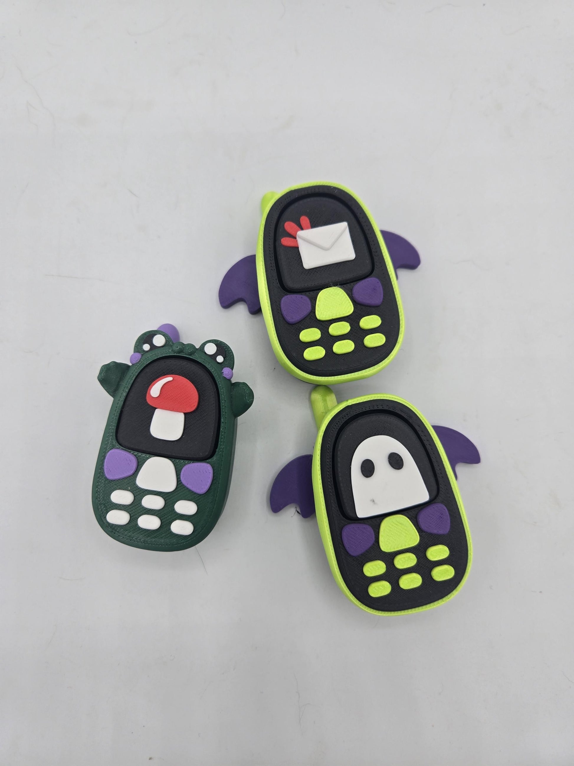 All three kawaii phone clicker styles together — frog with mushroom screen, bat with envelope screen, and bat with ghost screen, 3D printed