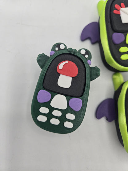 Kawaii frog phone clicker close-up — dark green froggy phone with red mushroom screen and purple accents, bat phones beside it, 3D printed