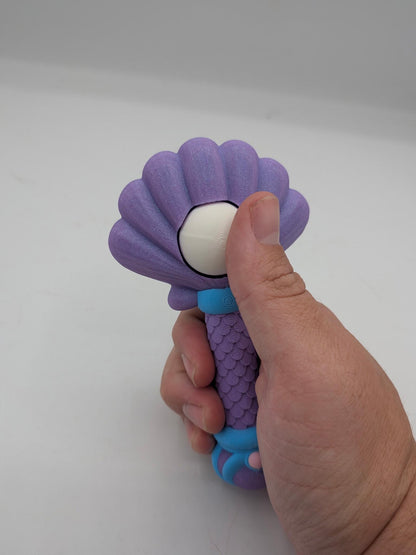 Hand holding the purple mermaid wand fidget with a thumb pressing the pearl clicker under the shell top