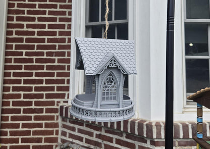 Gothic church bird feeder hanging outdoors against a brick wall — shingled roof and cathedral window