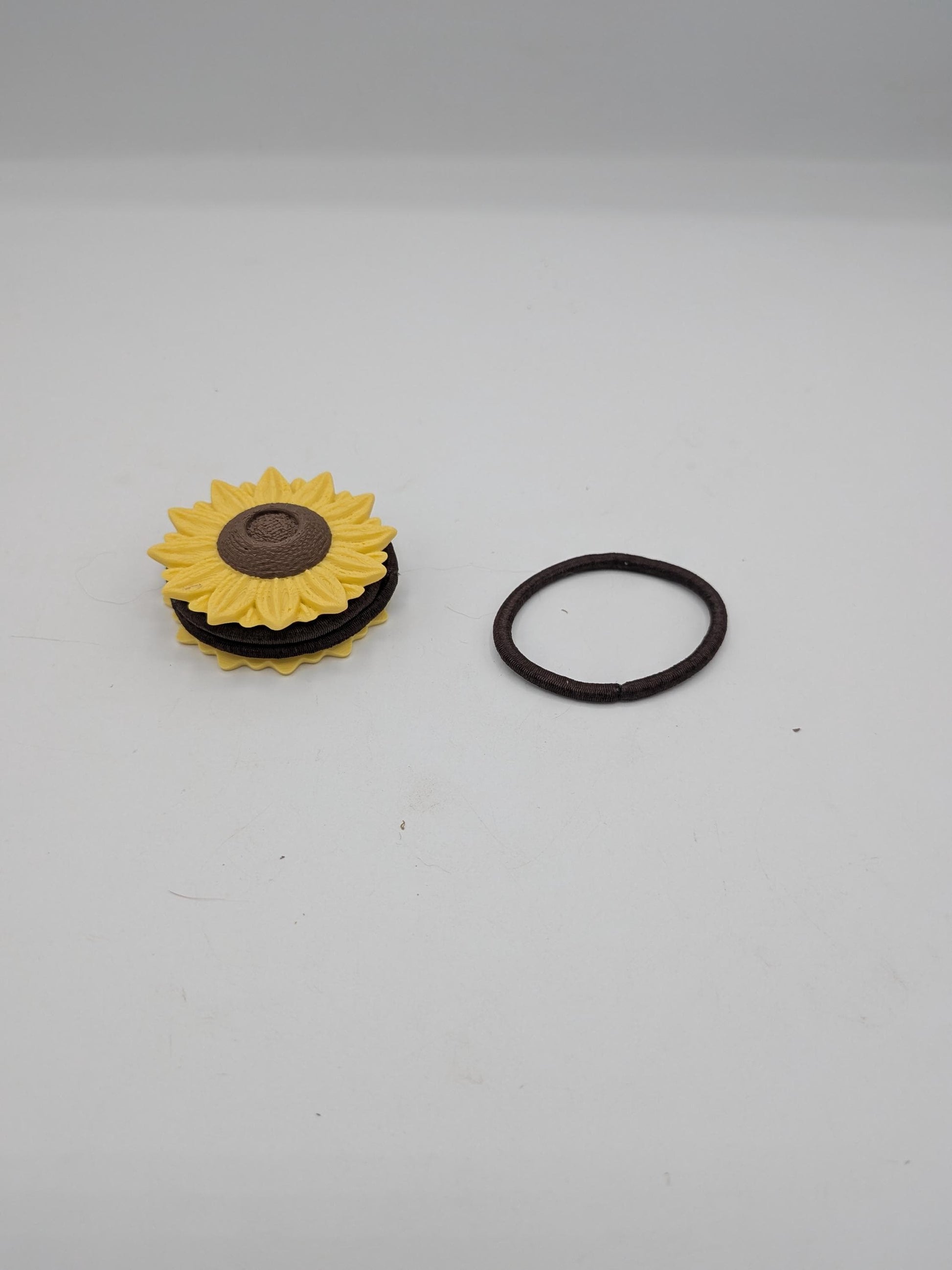 Sunflower hair tie holder 3D print with yellow petals and brown center, shown at an angle beside a brown elastic hair tie