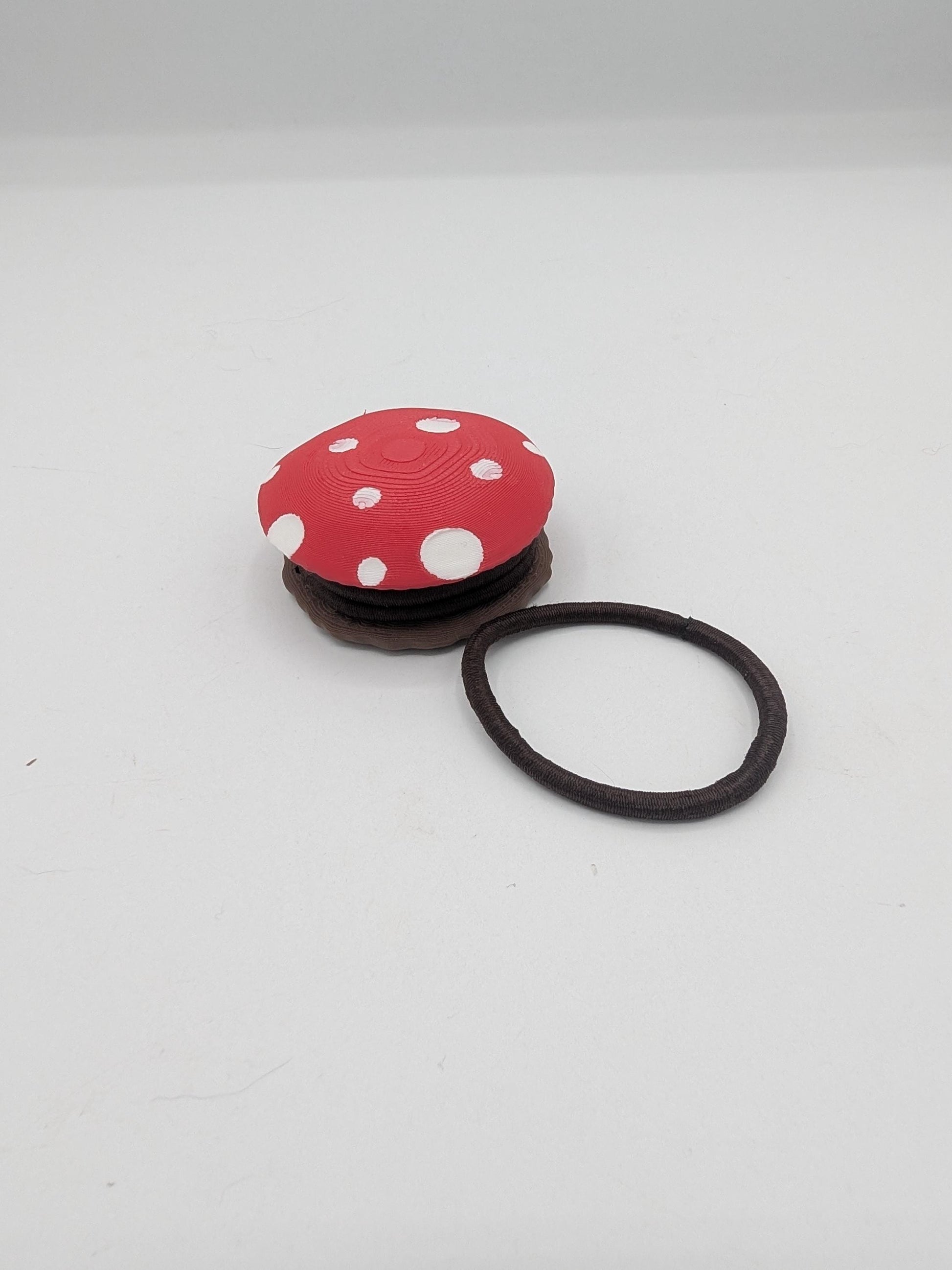 Mushroom hair tie holder 3D print with red and white spotted cap, angled view beside a brown elastic hair tie