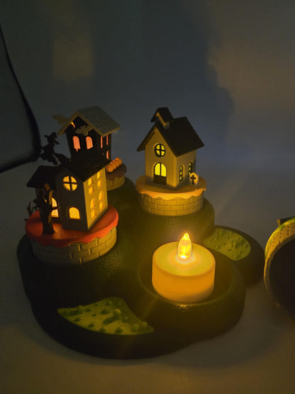 Halloween village at night with a flameless LED tealight glowing in an open base slot beside the lit haunted houses