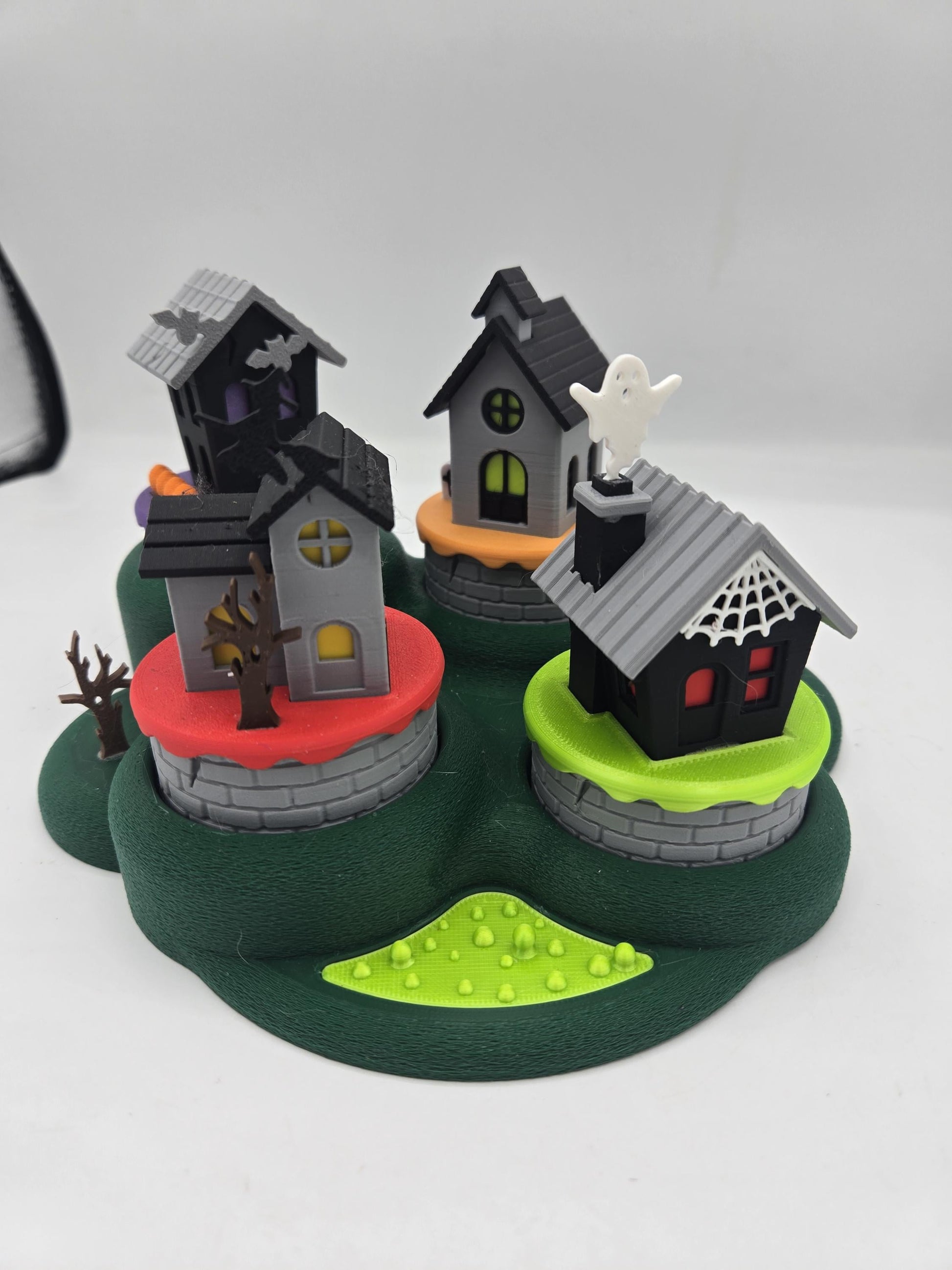 Halloween mini village tealight decoration — four 3D printed haunted houses with bats, a ghost, a witch, and spiderweb details on a green base, daylight view