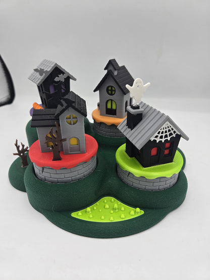 Halloween mini village tealight decoration — four 3D printed haunted houses with bats, a ghost, a witch, and spiderweb details on a green base, daylight view