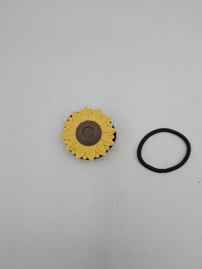 Top-down view of the sunflower hair tie holder next to a brown elastic hair tie, showing the flower cap that keeps stored ties in place
