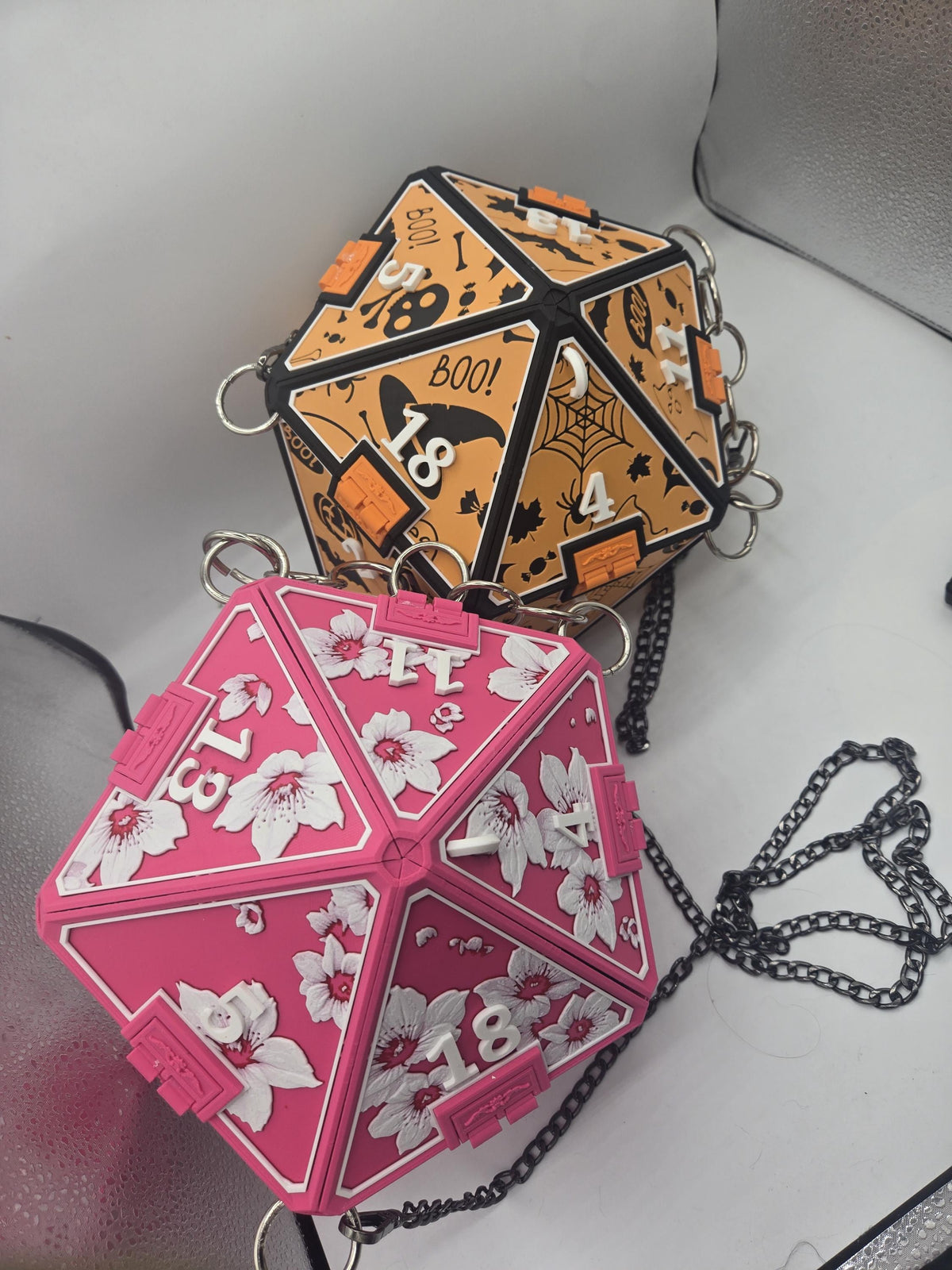 D20 Dice Shaped Purse 3D print, two color designs side by side: orange Halloween panels with ghosts, bats and BOO text, and pink cherry blossom panels, each with a black chain strap