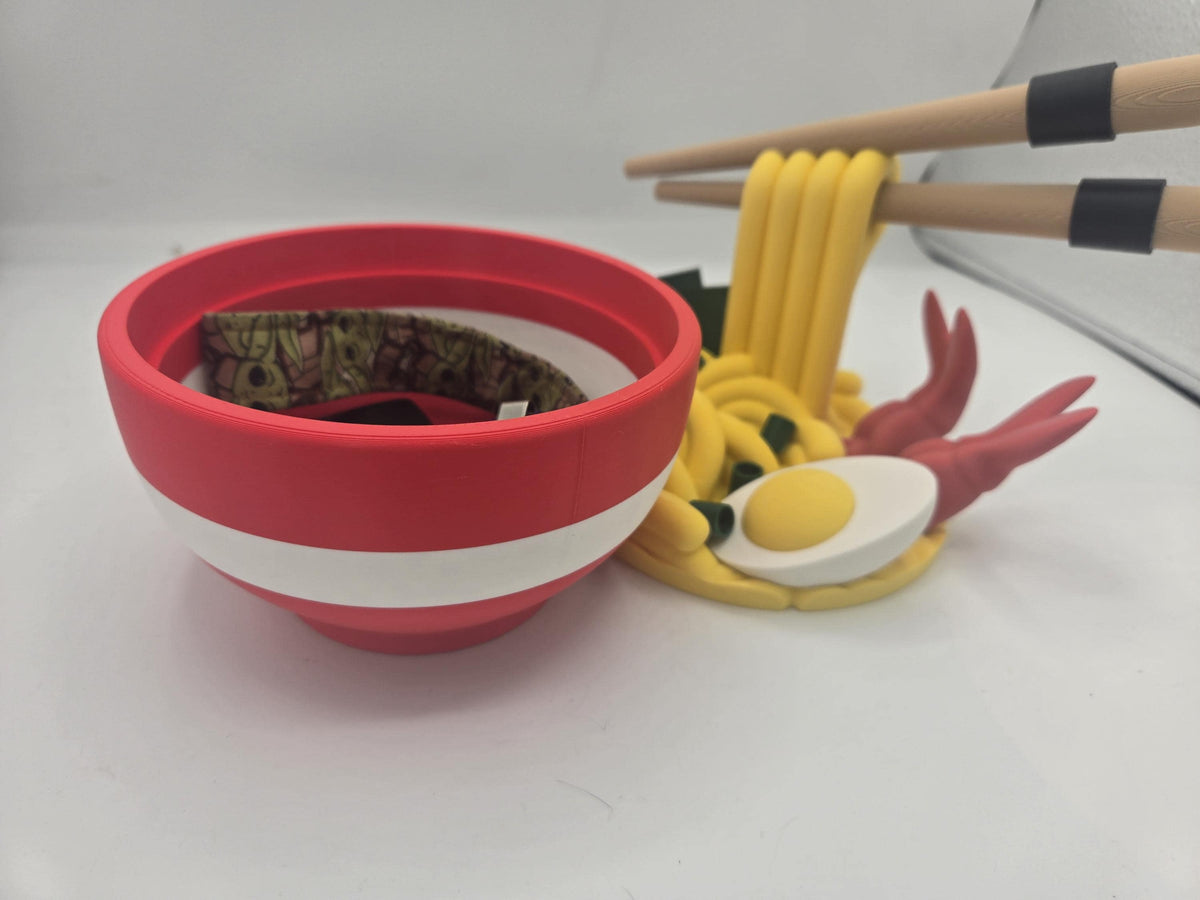 Ramen Bowl Display and Stash 3D print red bowl holding a small fabric wrist strap inside, noodle topper with egg and shrimp behind