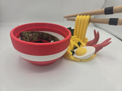 Ramen Bowl Display and Stash 3D print red bowl holding a small fabric wrist strap inside, noodle topper with egg and shrimp behind