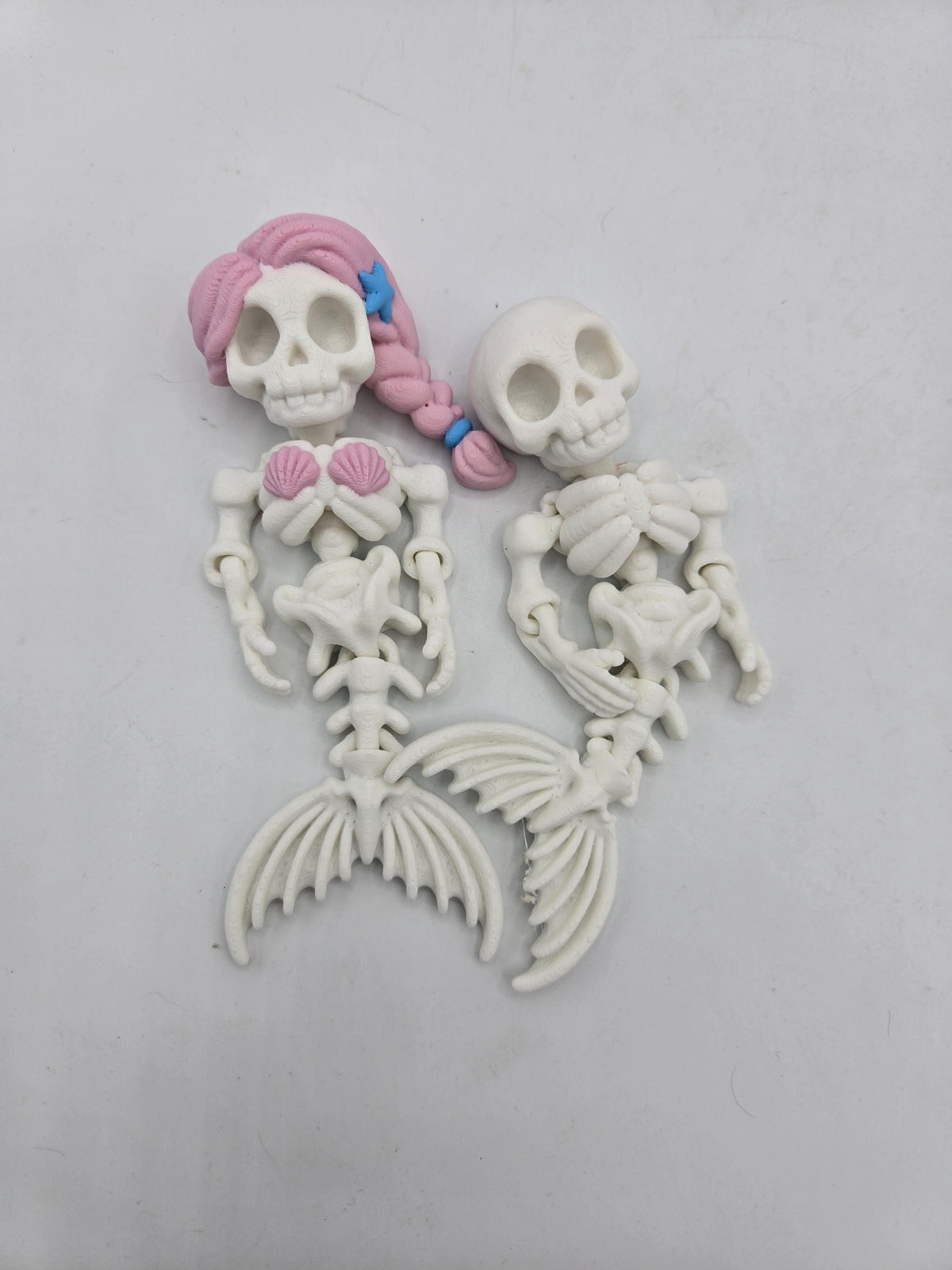 Flexi Mermaid Skeleton 3D print in white, both styles side by side: Hair + Clam Shells version with pink braided hair, blue starfish clip and pink clamshell top beside the plain skeleton mermaid