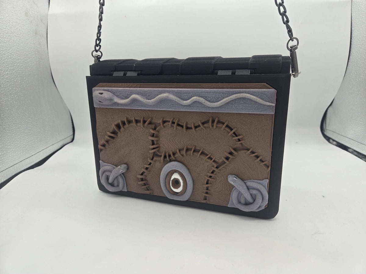 Storybound Book Themed Purse 3D print with stitched brown spellbook front panel featuring a single eye, coiled grey snakes and black book spine, hanging from a gunmetal chain strap